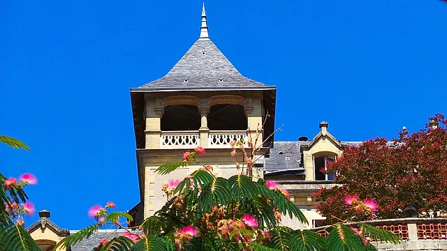 A slate-roofed tower with a balustraded loggia in Montignac, framed by pink mimosa flowers against a deep blue sky